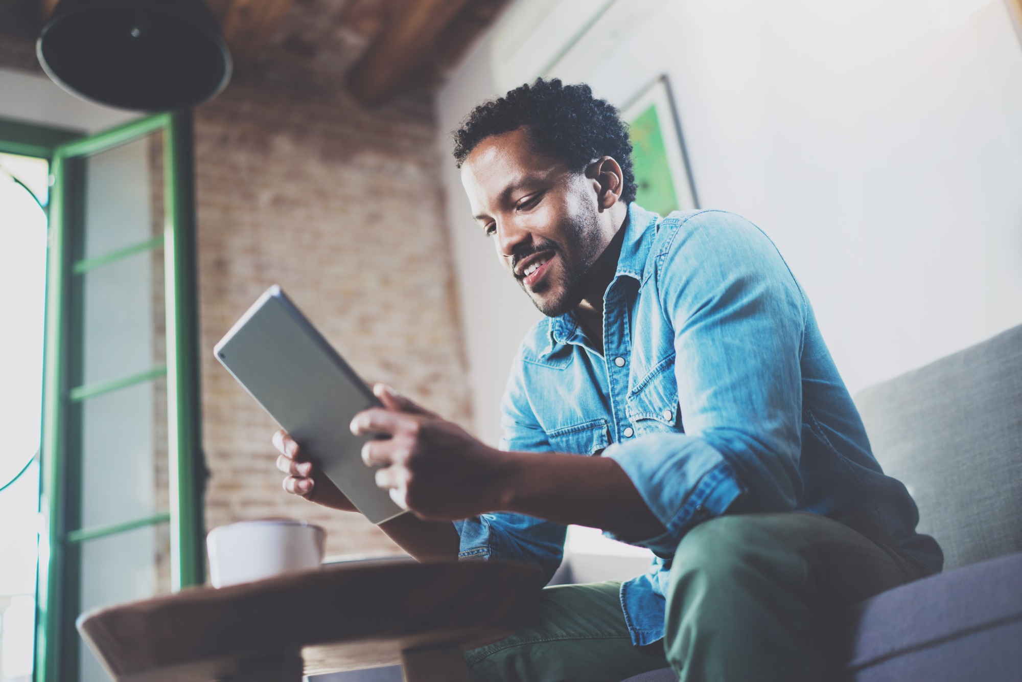 Bearded smiling African man using tablet for video conversation while relaxing on sofa